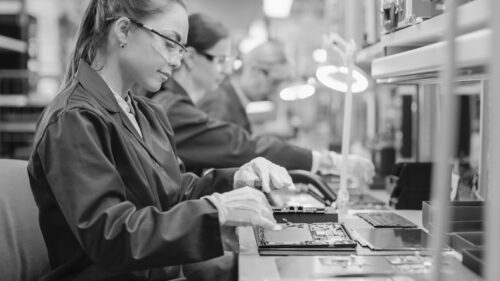 marketing for electronics manufacturing. Female Electronics Factory Worker in Blue Work Coat and Protective Glasses is Assembling Laptop's Motherboard with a Screwdriver. High Tech Factory Facility with Multiple Employees.